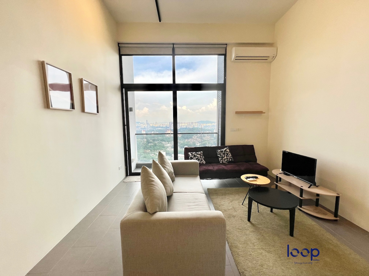 A bright living area features a spacious sofa facing large glass doors leading to a balcony. A small coffee table sits in front of a light-colored couch, while two framed pictures hang on the wall. Views of the city skyline are visible through the windows.