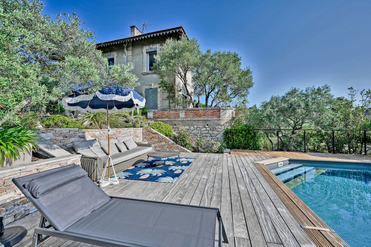 A spacious outdoor area features a wooden deck surrounding a rectangular pool. A sunbed is positioned beside the pool, with a decorative umbrella providing shade. Olive trees and greenery add to the serene atmosphere, enhancing the view of the historic house in the background.