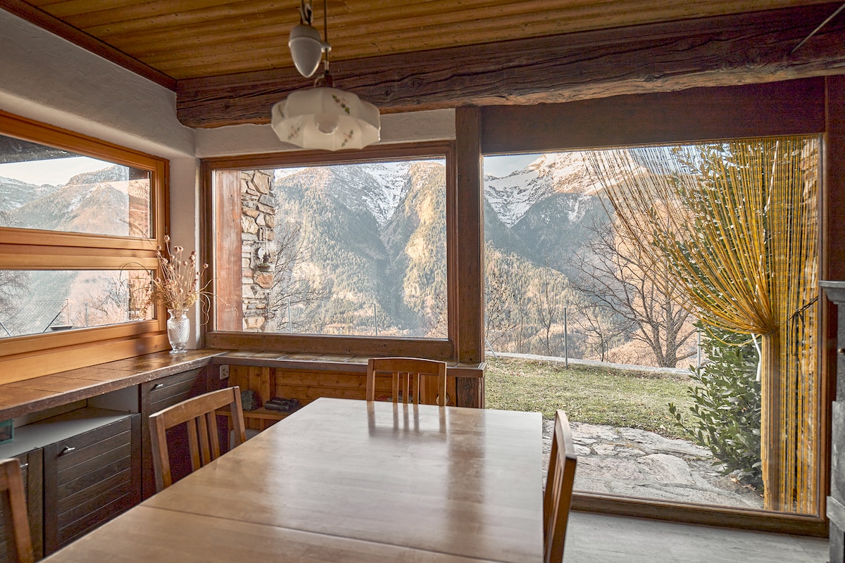 A wooden dining table is positioned near large windows, providing panoramic views of surrounding mountains and nature. Soft light filters through the windows, while a simple vase adds a touch of decor to the space.