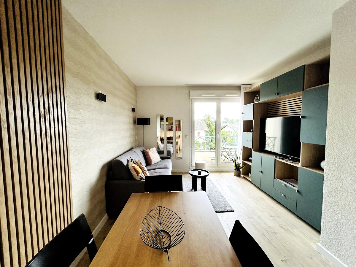 A bright living area features a comfortable grey sofa adorned with colorful cushions, positioned next to a sleek television unit. A wooden dining table is set for four, accompanied by black chairs. Large windows allow natural light to illuminate the space, offering a view of the outdoor balcony.