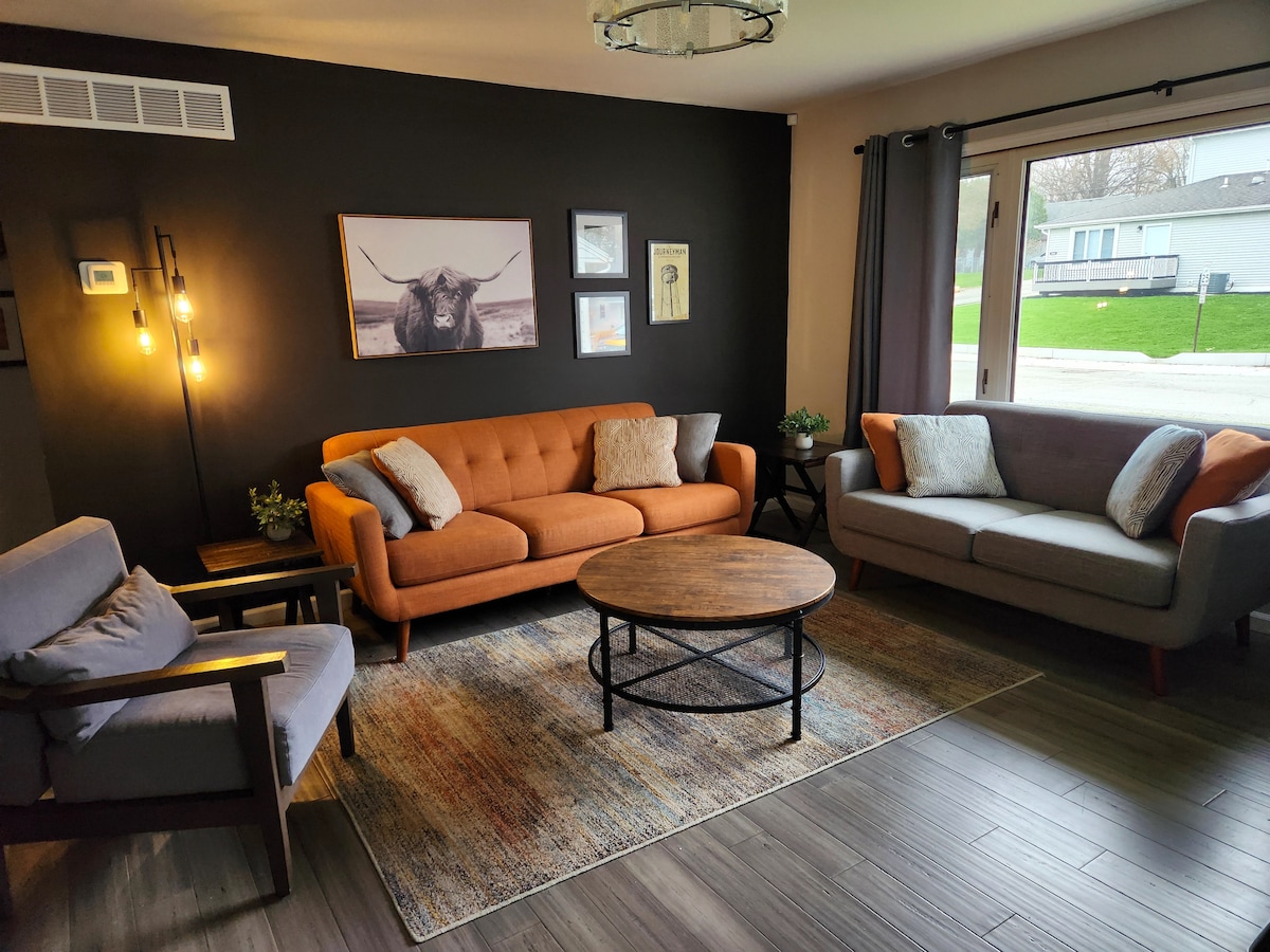 A cozy living area features a circular coffee table centered on a textured rug. An orange sofa and gray armchairs are positioned around the table. Artwork of a bull adorns the dark wall behind. Natural light filters through sliding glass doors leading to the outdoor space.