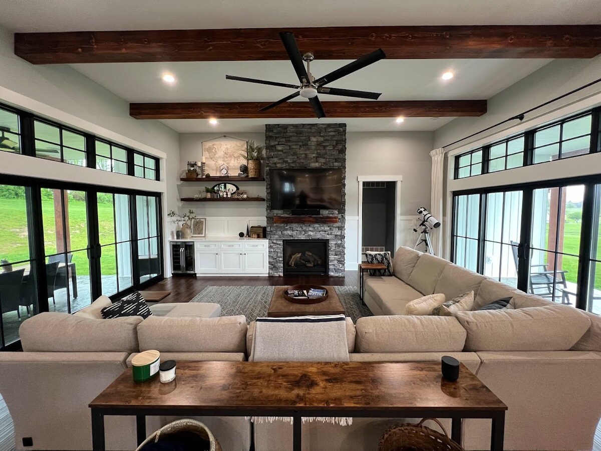 A spacious living room features large glass doors that invite natural light, complemented by dark wood beams overhead. A comfortable sectional sofa encircles a low coffee table, while a stone fireplace is centrally located against a feature wall. Decor and a television enhance the cozy setting.