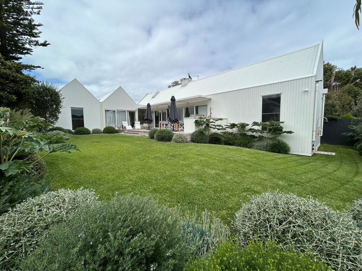 The exterior of the beach house is presented, featuring a well-manicured lawn and contemporary white structures. Large windows allow natural light to enter the living spaces. Outdoor seating areas are available, complemented by umbrellas for shade. Lush greenery surrounds the property, enhancing the tranquil setting.