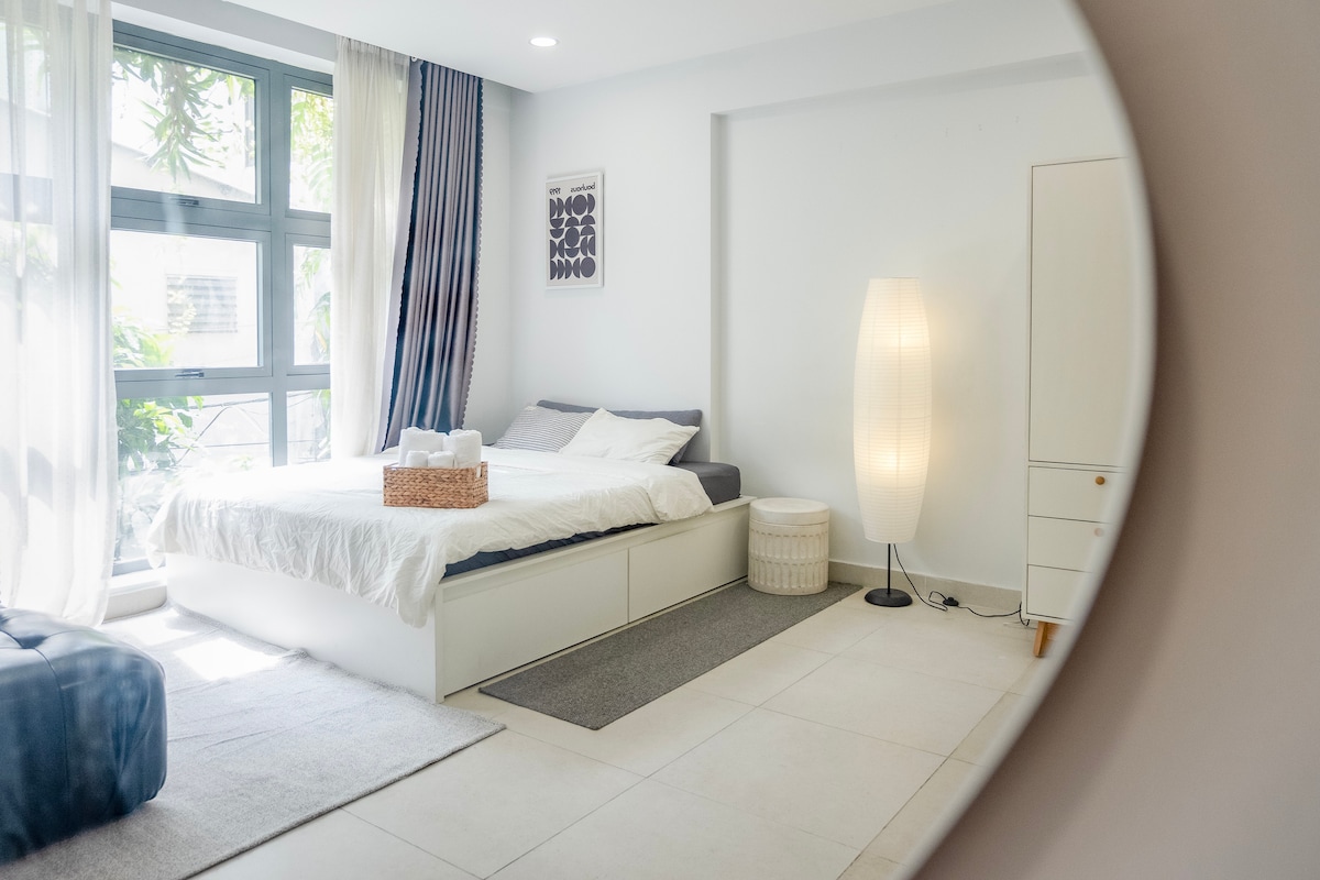 A light-filled room is visible, featuring a spacious bed with neutral bedding. A wicker basket sits at the foot of the bed. A soft area rug is placed under a stylish lamp, and a large window offers a view of greenery outside.