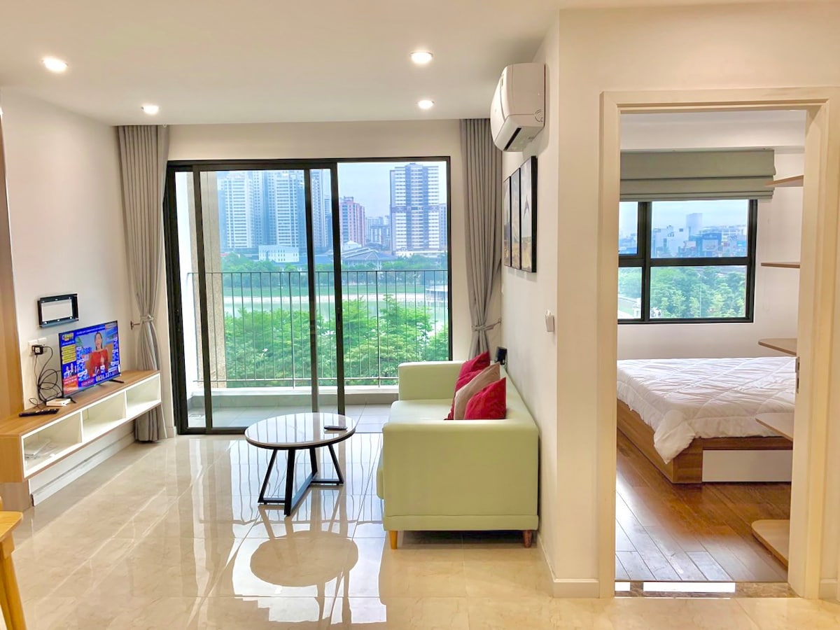 A bright and airy living area is featured with a green sofa and a coffee table, providing a comfortable space for relaxation. A large window showcases views of the city skyline and greenery outside. The bedroom is visible in the background, separated by an open door.