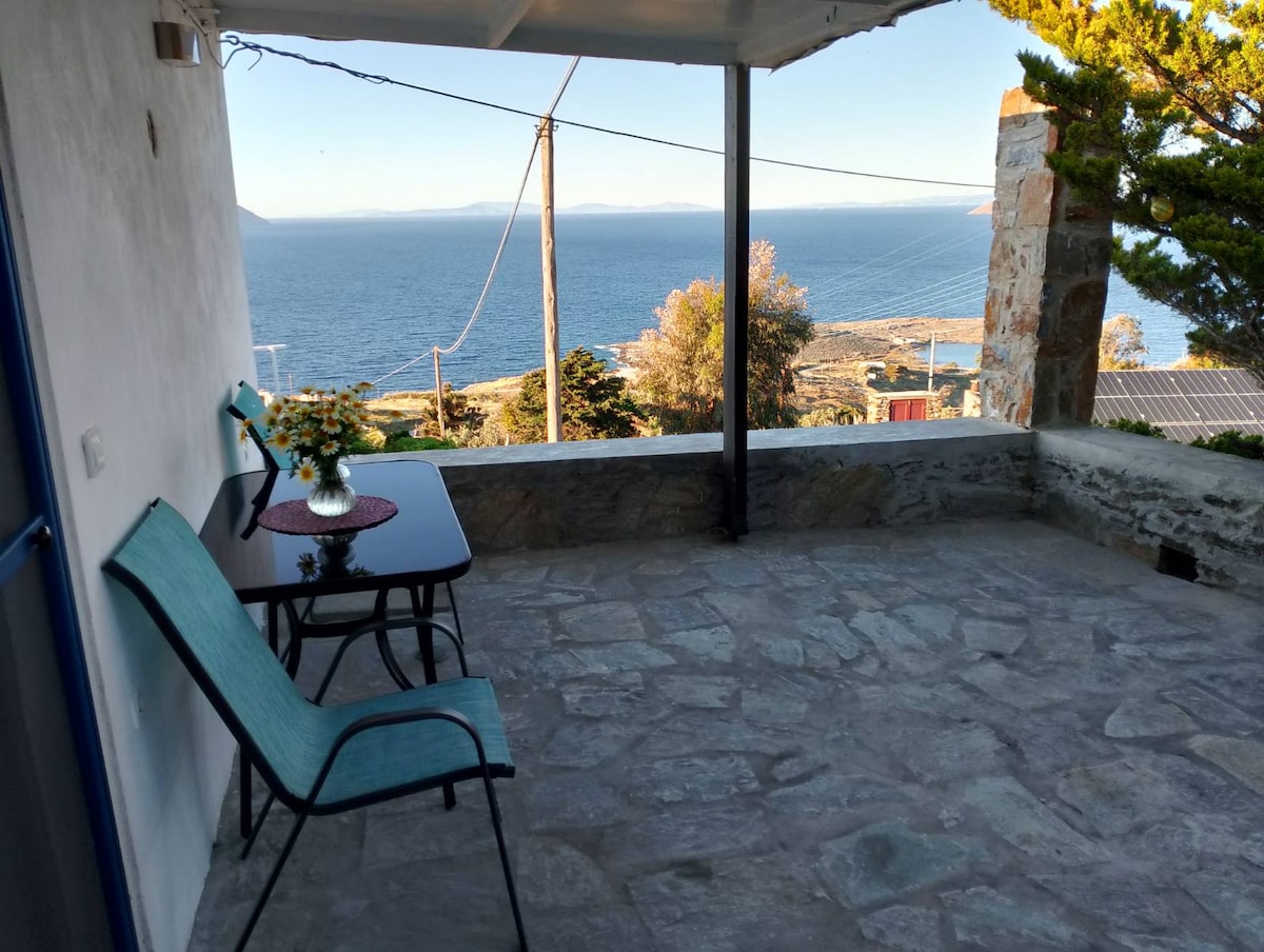 A covered outdoor terrace displays a small glass table accompanied by two chairs. The stone flooring enhances the space, while the backdrop features a stunning view of the sea and distant hills, creating a serene environment for relaxation.