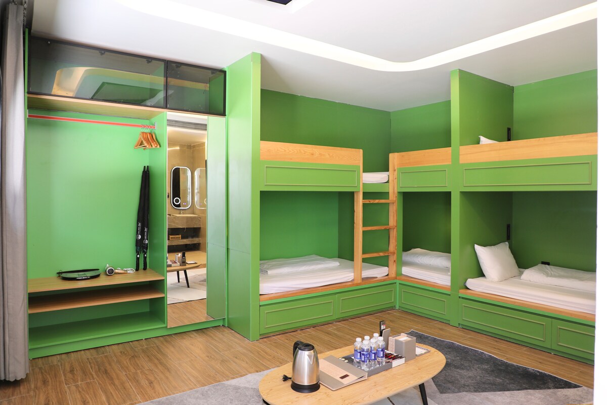 A vibrant green room features two sets of wooden bunk beds, providing ample sleeping space. A small table with bottles of water and a kettle sits in the center. A corner showcases a closet with hangers, and a glass partition allows a view of the bathroom.