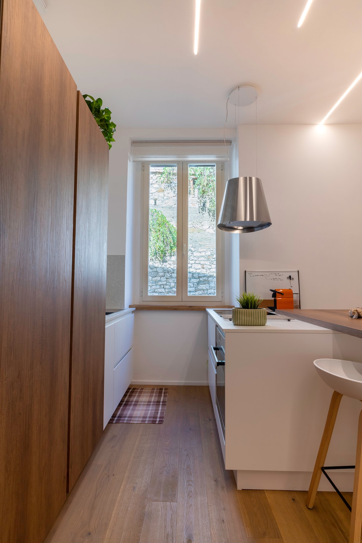 A modern kitchen space features white cabinetry and a wooden accent wall. Natural light floods the area through a window that overlooks greenery. A metal pendant light hangs above a compact countertop, while a small plant adds a touch of color to the setting.