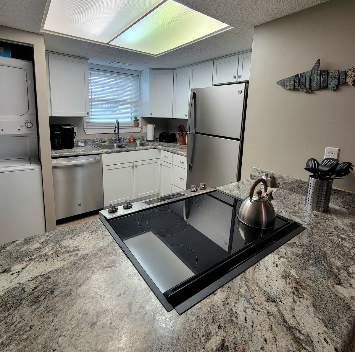 The kitchen is equipped with modern appliances, including a refrigerator, dishwasher, and a stove with an integrated cooktop. A granite countertop provides ample space for meal preparation, while cabinets and a coffee maker are visible in the background. Natural light brightens the area through a window.