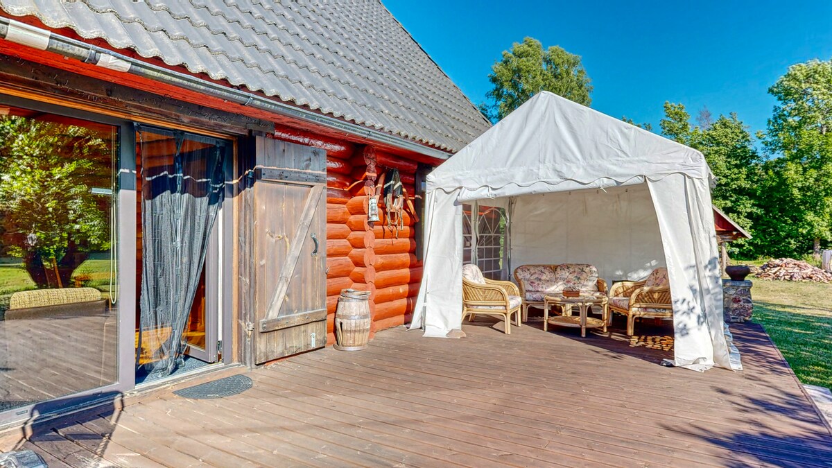 A covered outdoor seating area is situated on a wooden deck, featuring wicker furniture arranged for relaxation. Nearby, the rustic wooden exterior of the house is visible, complemented by clear blue skies and greenery in the background.