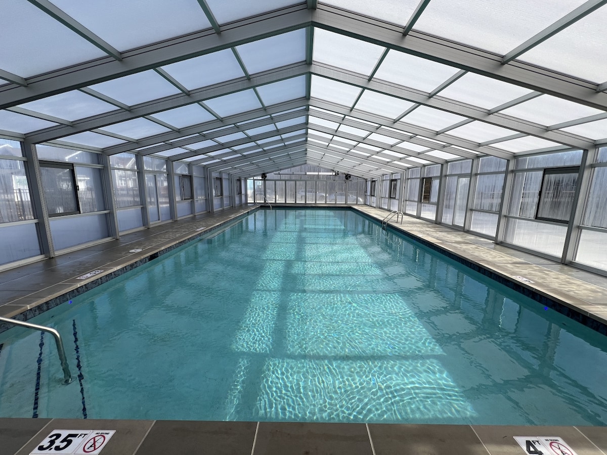An indoor pool area is seen, featuring a spacious swimming pool with clear water. Large windows allow natural light to illuminate the space, highlighting the clean lines of the modern design. Ladders are positioned at either end, and depth markers are visible along the edge.
