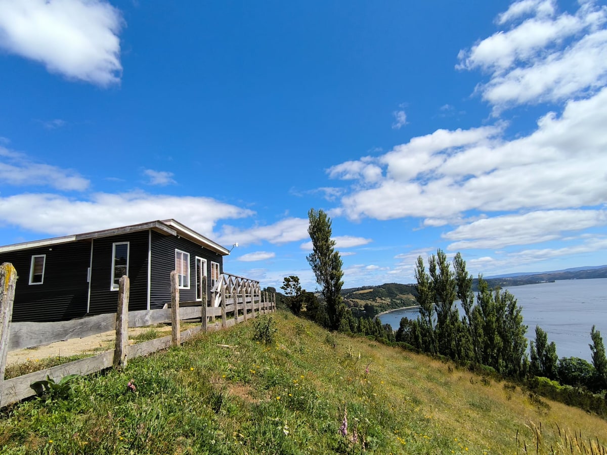 A black wooden cabin sits atop a hill, with expansive views of a tranquil body of water and a clear blue sky. The surrounding landscape features green grass and scattered wildflowers, and a wooden fence lines the pathway leading to the cabin.