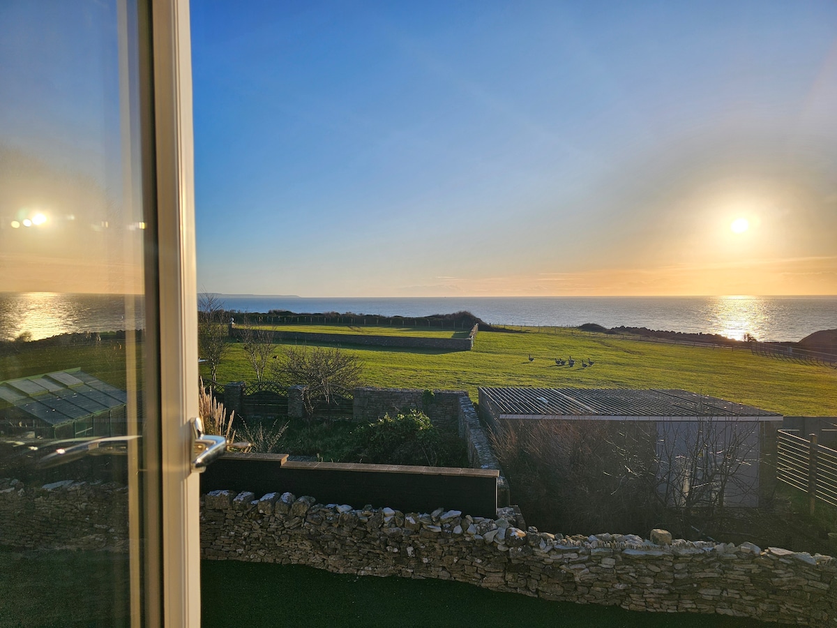 A captivating view extends from the window, revealing a sunlit sea blending into the horizon. Lush green fields are framed by a stone wall, while the golden light of sunset casts a warm glow over the tranquil landscape.
