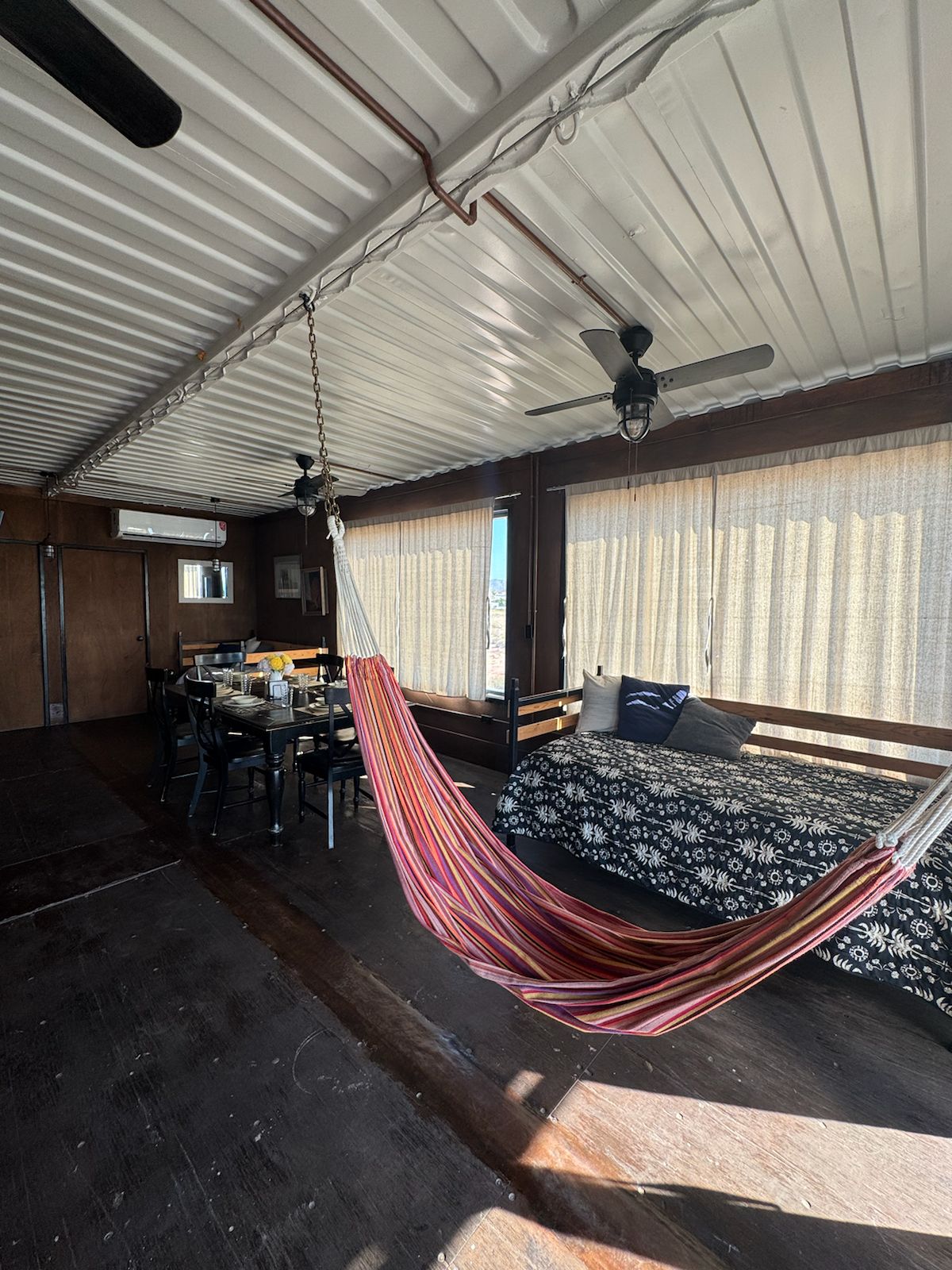 The interior space features a colorful striped hammock suspended from the ceiling, with light streaming through sheer curtains. A dining table is positioned nearby alongside a cozy seating area, adorned with patterned textiles, offering a relaxed and inviting space for guests.
