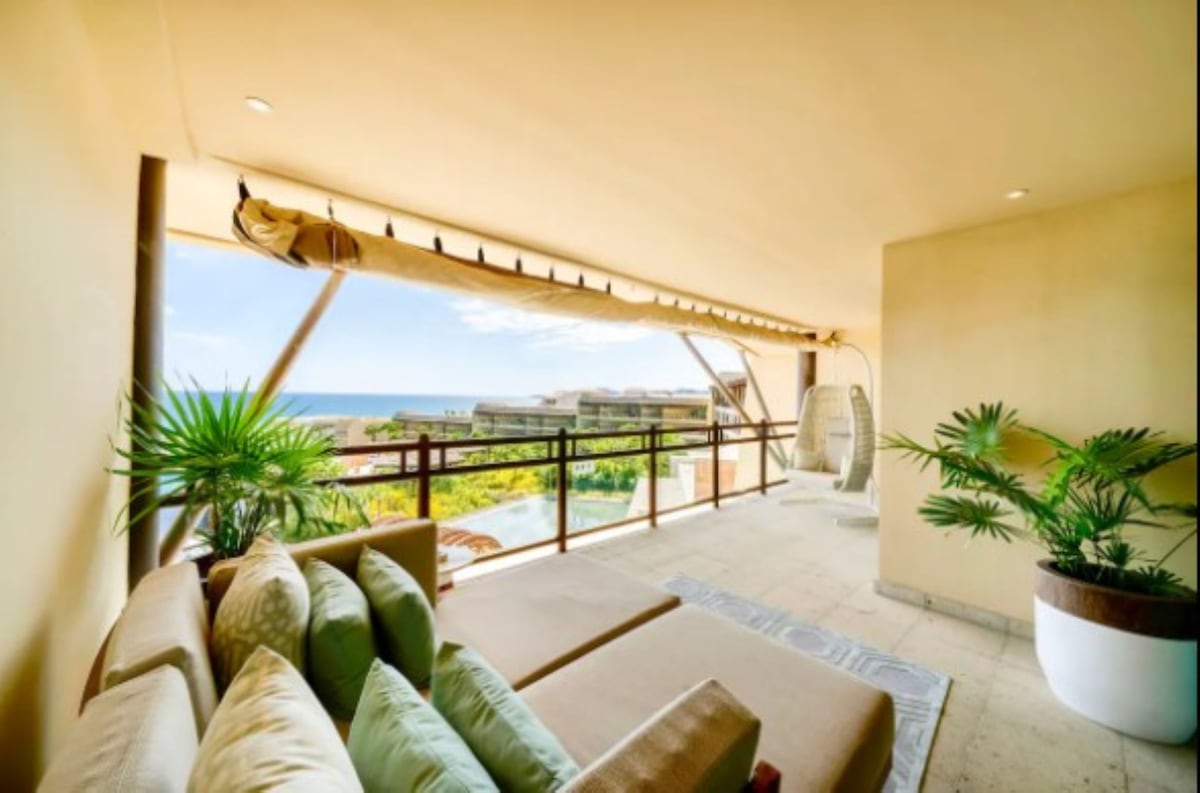 A spacious balcony is presented with a modern seating arrangement featuring soft cushions. Potted plants add greenery, and a distant view of the ocean can be seen through the railing. Natural light enhances the warm tones of the space.