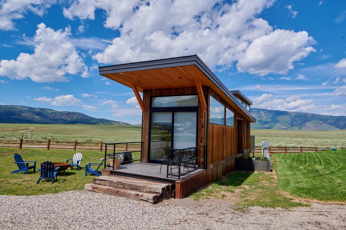A modern tiny home is set against a scenic landscape, featuring large windows that invite natural light. An outdoor seating area includes several chairs positioned on a deck, offering views of the surrounding greenery and mountains. The exterior combines wood and glass elements for a contemporary feel.