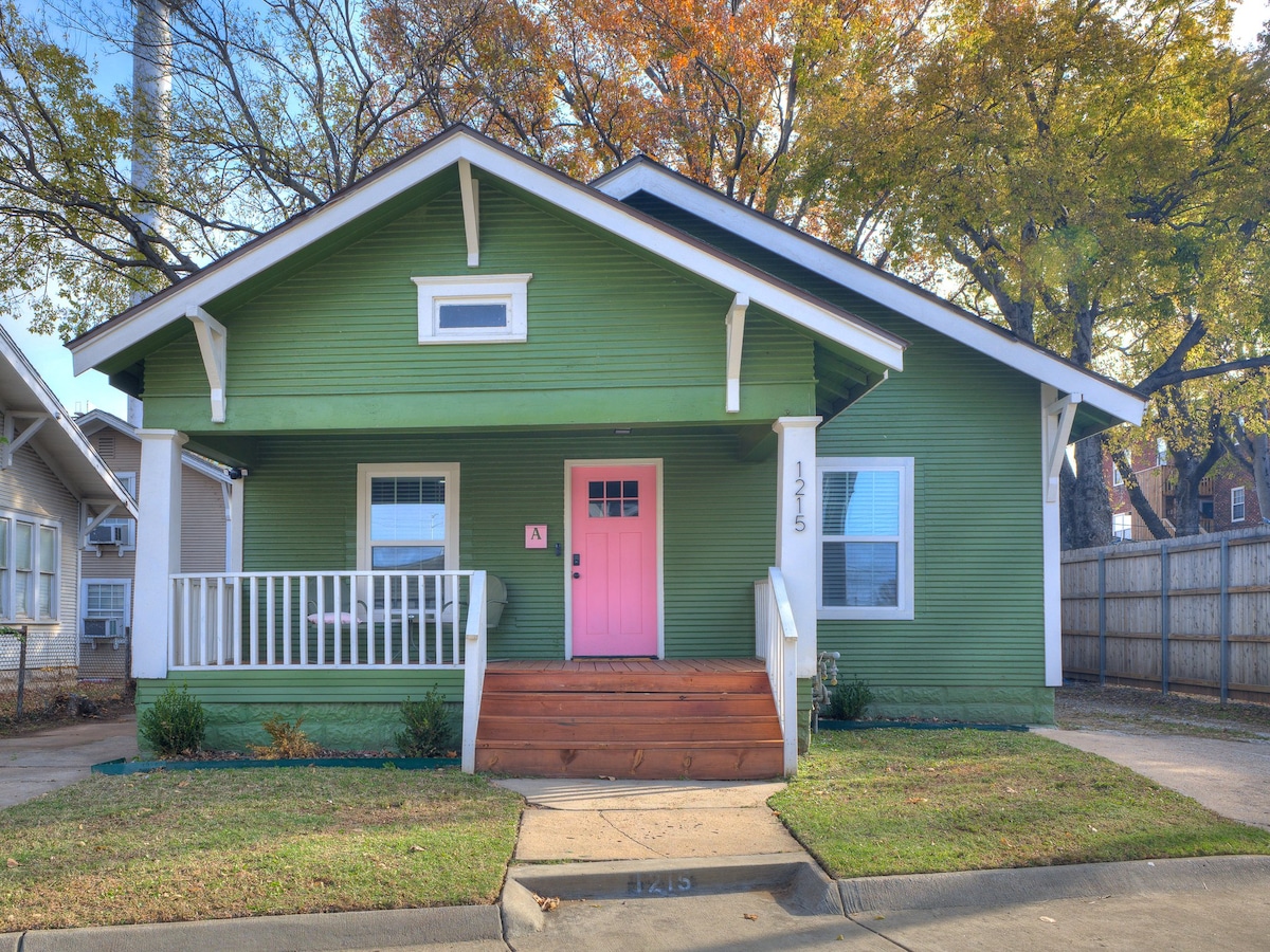 The exterior of the duplex features a vibrant green facade, complemented by a bright pink front door. A welcoming porch is accessed via wooden steps, framed by white railings. Large windows allow natural light, and the surrounding area includes well-maintained grass and seasonal trees.