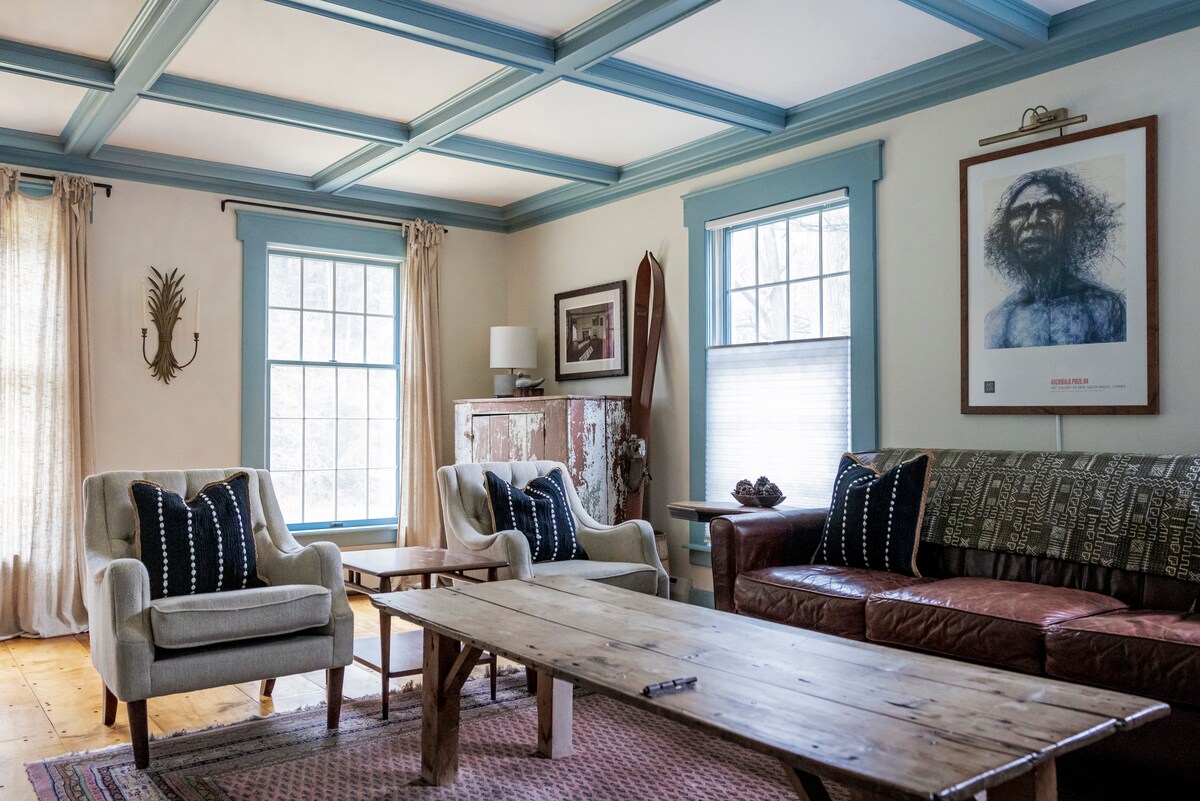 A formal living room features a coffered ceiling with blue accents. Two comfortable chairs accompanied by a textured sofa provide ample seating. A rustic coffee table is centered on a patterned rug, while natural light filters in through two windows with blue trim.