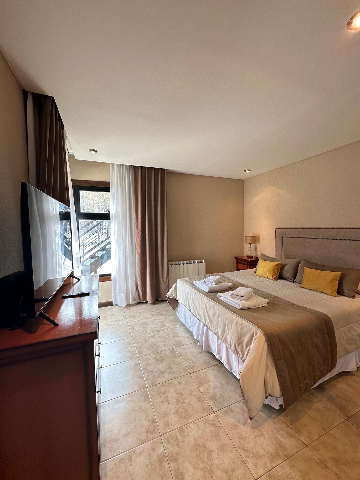 A light-filled bedroom includes a spacious bed adorned with neutral-toned linens and three accent pillows. A television is positioned on a wooden dresser. Large windows reveal natural light and are flanked by soft curtains, enhancing the room's overall brightness.