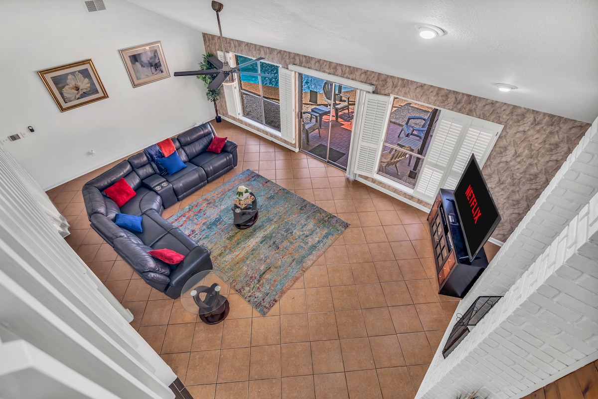 A spacious living area showcases a large sectional sofa accented with vibrant cushions and a central coffee table. Natural light pours in through multiple windows, which are framed by stylish shutters. A television is positioned for easy viewing, complementing the inviting layout.