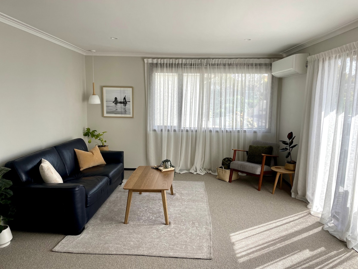 A spacious living area features a dark leather sofa and a wooden coffee table on a soft area rug. Natural light filters through sheer curtains, illuminating a pair of accent chairs and a small side table. A potted plant adds a touch of greenery.