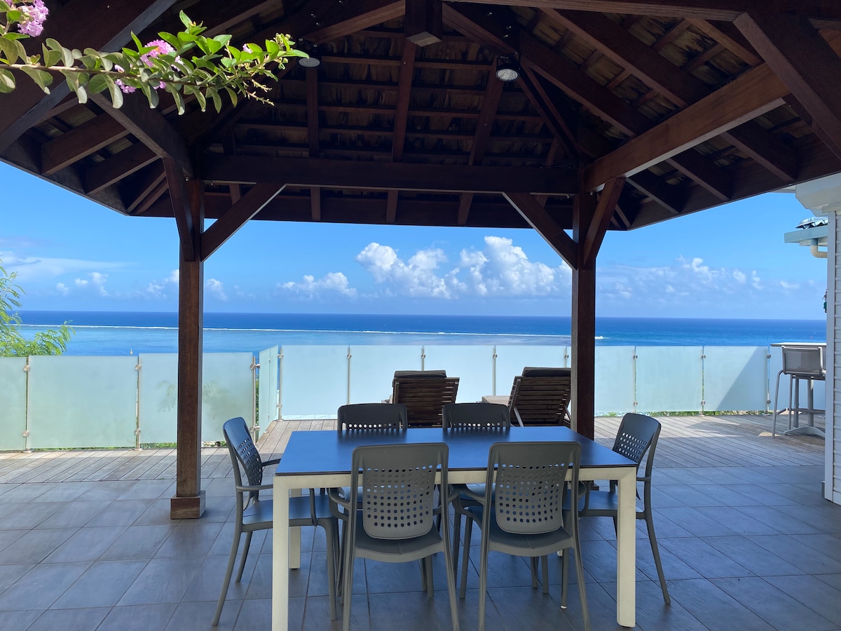 An outdoor dining area is situated under a wooden pavilion, featuring a long white table with gray chairs. The backdrop showcases a panoramic ocean view, with clear blue skies and scattered clouds visible beyond the railing. Lush green foliage frames the scene.