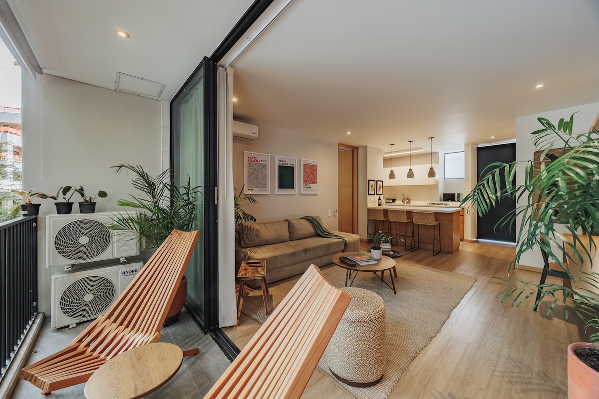 A cozy living space is depicted with a comfortable sofa facing a well-equipped kitchen. Two modern loungers are placed on a balcony, surrounded by green plants. Large glass doors allow natural light to fill the area, creating an open and inviting atmosphere.