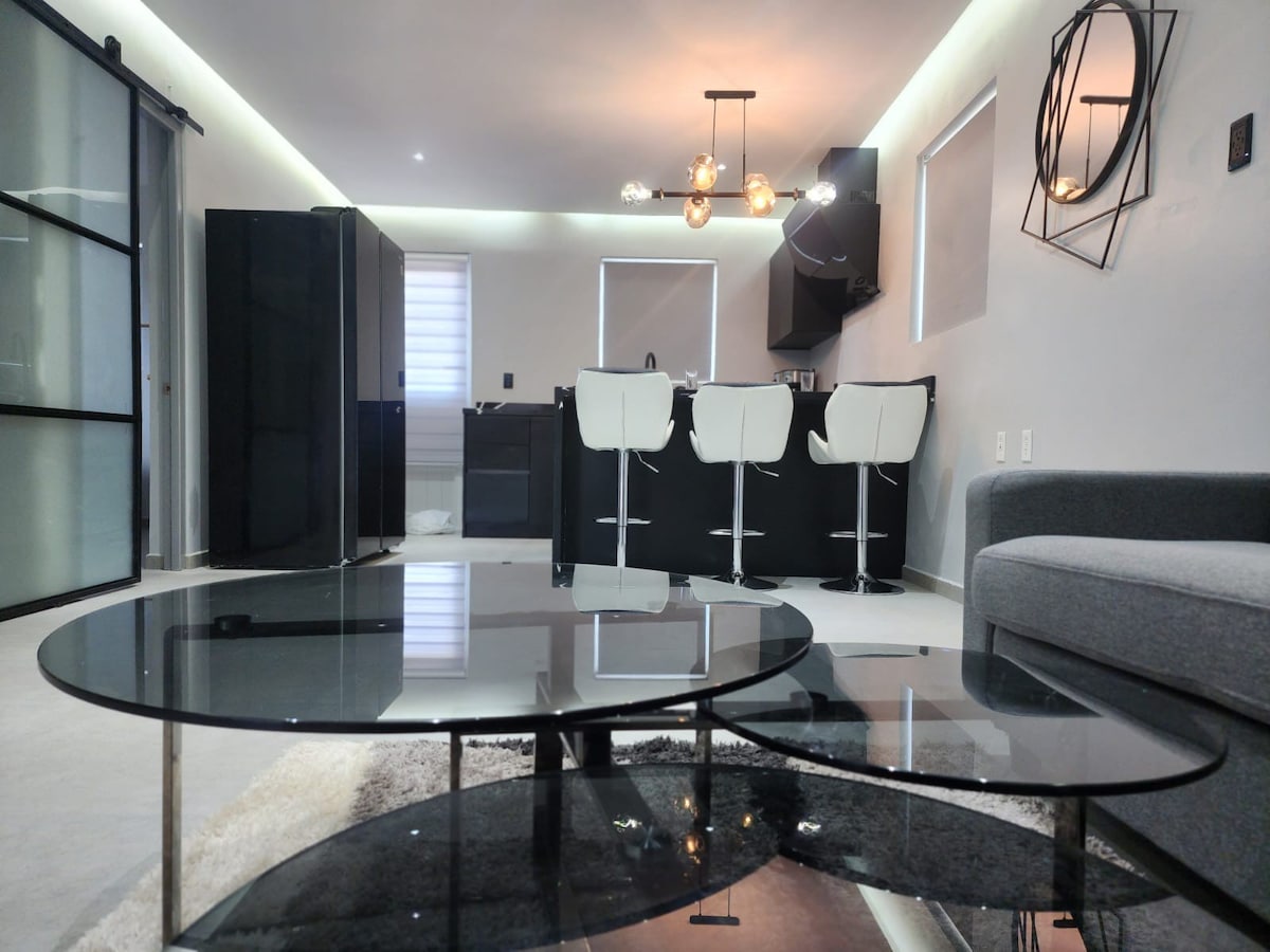A modern living space is showcased with sleek furniture. Two stylish tables with glass tops are positioned in front of a comfortable grey sofa. The open kitchen area features black cabinetry and a dining counter with three white bar stools illuminated by warm overhead lights.