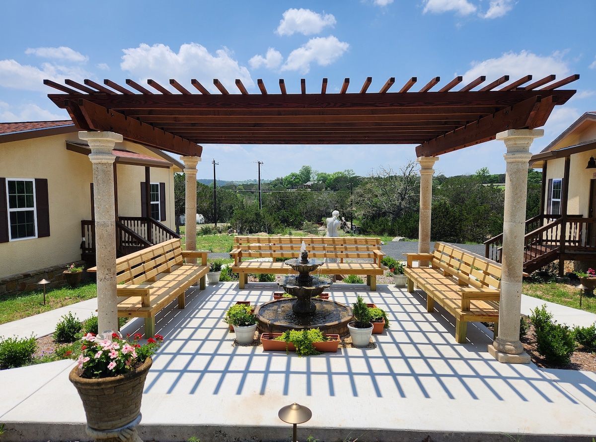 An outdoor courtyard features a wooden pergola supported by stone columns, providing shade over several wooden benches arranged around a central fountain. Potted plants are placed throughout the area, complementing the lush greenery visible in the background under a clear blue sky.