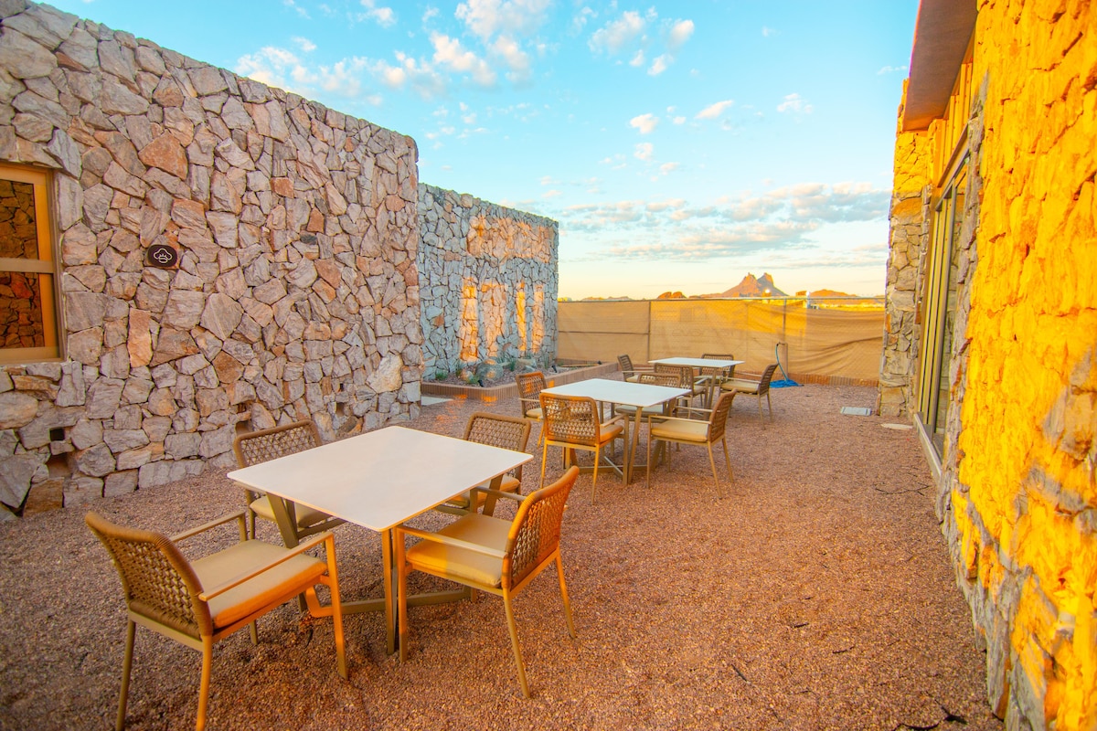 An outdoor dining area features multiple tables with light-colored surfaces and woven chairs. Stone walls made of natural materials create a rustic backdrop, while the soft light of sunset bathes the space in warm tones. Scenic views of the surrounding landscape can be seen in the distance.