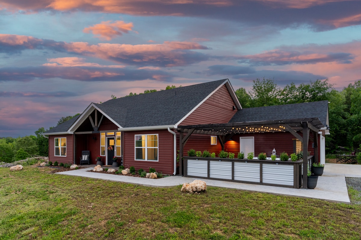A newly-built farmhouse is shown with a rich burgundy exterior and a covered patio area. The landscape features well-maintained greenery and rocks, complemented by a sunset sky filled with varying shades of pink and purple, creating a serene backdrop.