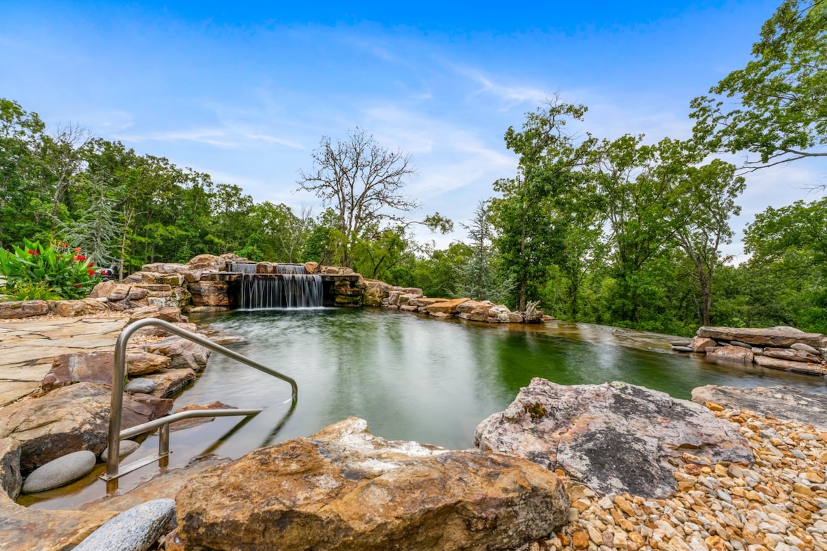 A natural swimming pond is surrounded by smooth stone. A small waterfall cascades over rocks, adding to the serene atmosphere. Lush greenery lines the pond, creating a secluded feel. The water reflects the blue sky, inviting a tranquil, relaxing experience.