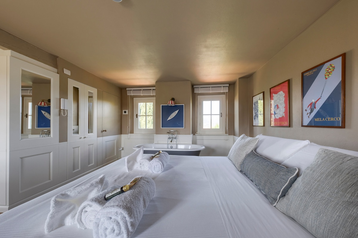 A spacious and elegantly decorated bedroom features a large bed adorned with white linens and accent pillows. Two windows allow natural light to fill the room, while art pieces hang on the walls. A stylish freestanding tub adds a touch of sophistication to the space.