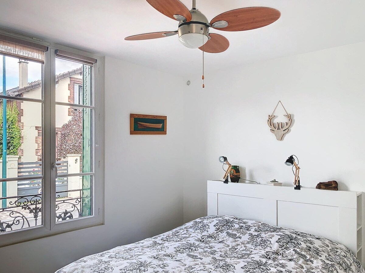 A bright bedroom features a spacious bed adorned with a floral-patterned duvet. Natural light floods the room through large windows with open views. A ceiling fan provides airflow, while a small bedside lamp and decorative elements add to the room's simplicity and comfort.