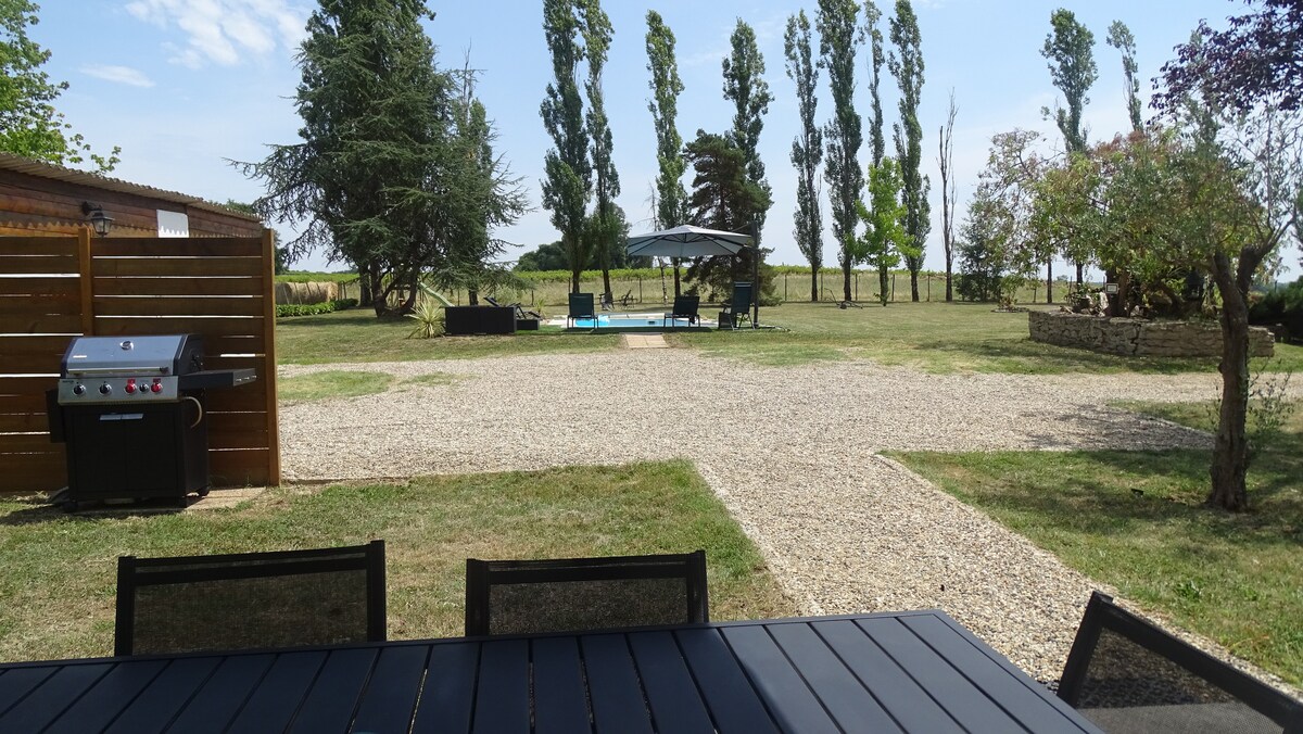 A spacious outdoor area is shown, featuring a gravel pathway leading to a blue pool surrounded by grass. Seating areas under umbrellas and shaded trees are visible. A barbecue grill and a dining table are positioned in the foreground, enhancing the outdoor living experience.