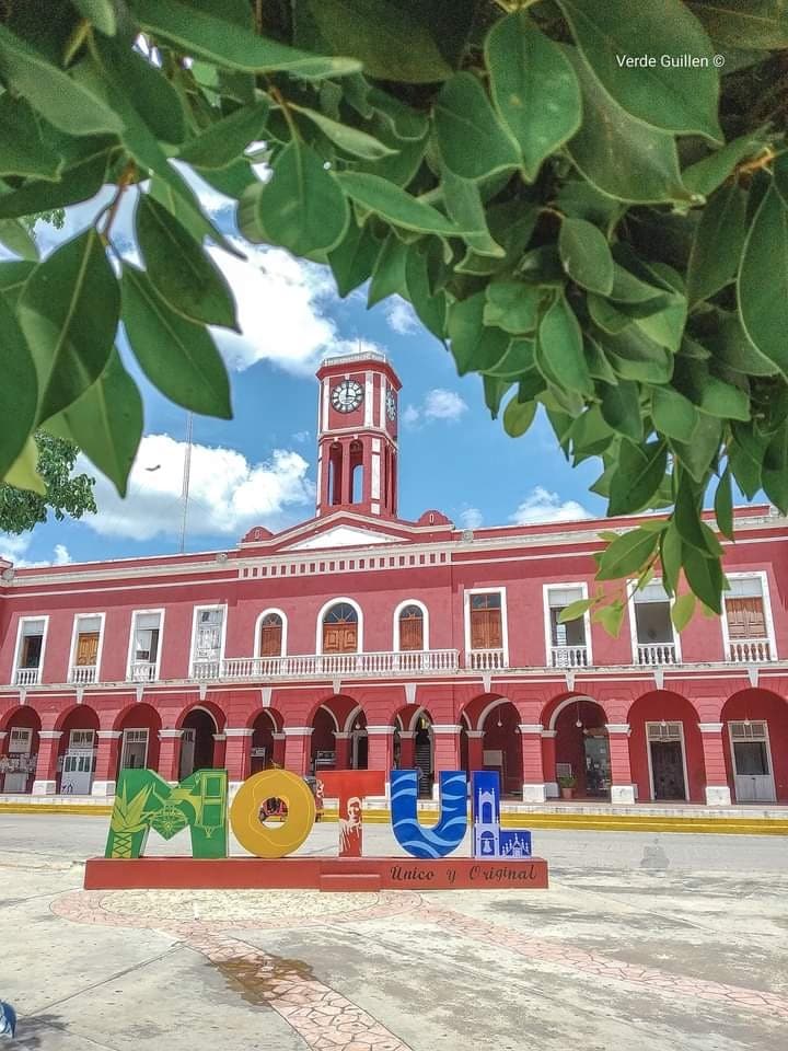 A vibrant building in a rich red color is featured, showcasing a clock tower at its center. In the foreground, colorful letters spell out the location's name, framed by lush green leaves from nearby trees, contributing to a lively community atmosphere.