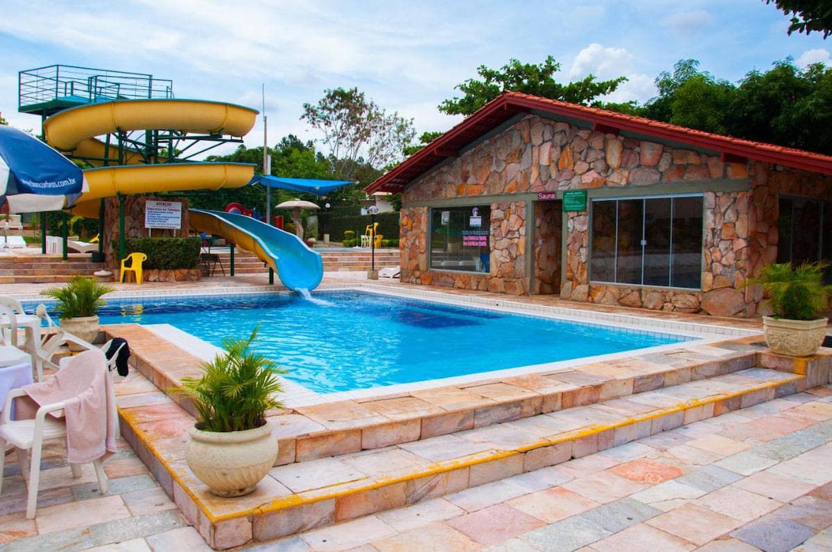 A vibrant swimming pool area is depicted, featuring a large water slide adjacent to a stone building. Tables and chairs are arranged nearby, with greenery accentuating the space. The pool's clear water invites relaxation, and the surrounding trees provide shade.