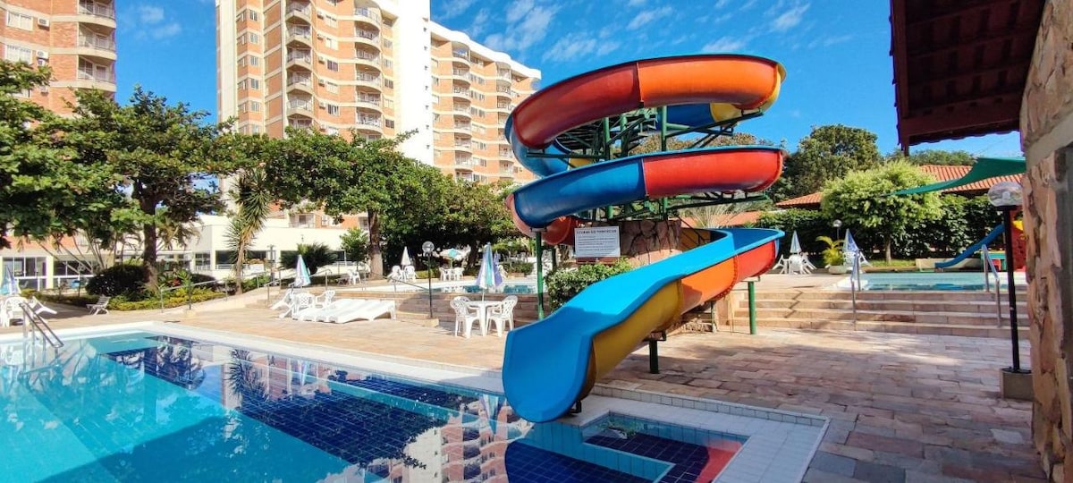 A vibrant water slide is positioned beside a clear pool, offering fun for guests. Surrounding the pool area, multiple lounge chairs are placed under palm trees, with a backdrop of the condominium's buildings. Lush greenery enhances the inviting outdoor space.