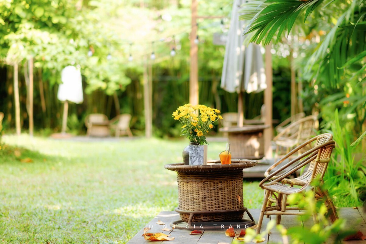 A serene garden setting is presented, featuring wicker chairs arranged around a circular table. A vase of yellow flowers rests on the table, accompanied by a drink. Lush greenery and outdoor umbrellas create a relaxed atmosphere in the inviting space.