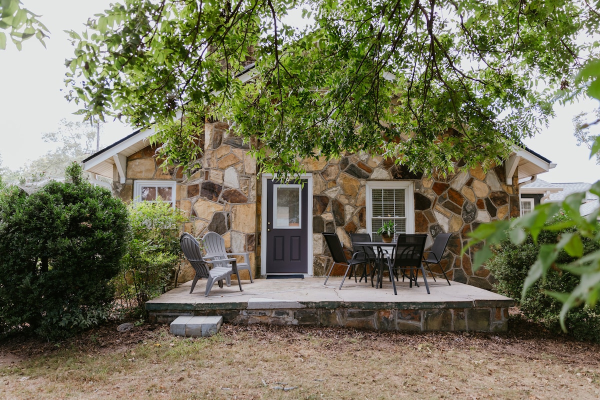 The exterior of the stone cottage is revealed, framed by lush greenery. A welcoming porch area features a dining table surrounded by chairs. Two Adirondack chairs are placed to the side, providing a cozy spot for relaxation. The door to the cottage stands invitingly ajar.