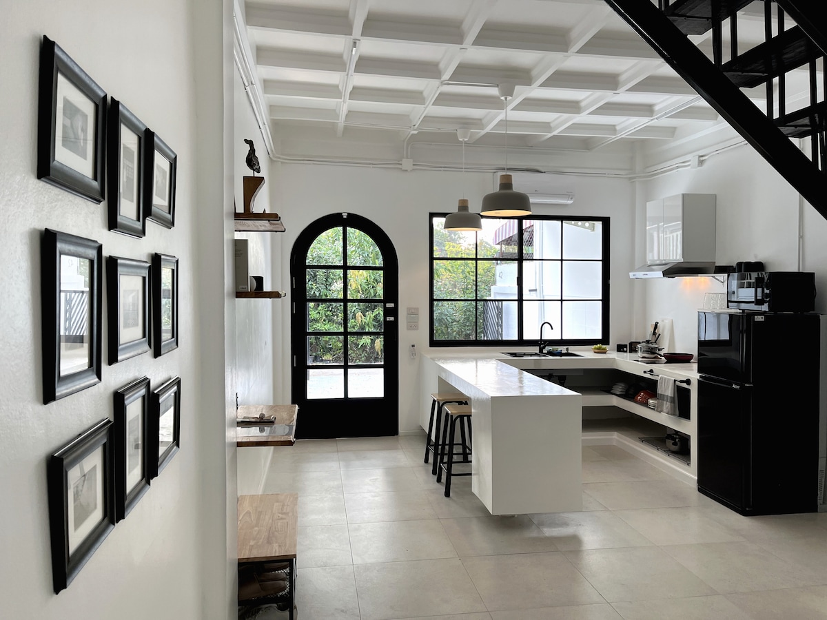 A modern kitchen area features an island with seating, sleek cabinetry, and a black refrigerator. Sunlight streams through a large arched window, illuminating the space and offering views of greenery outside. Framed photographs adorn the nearby wall, adding a touch of character.