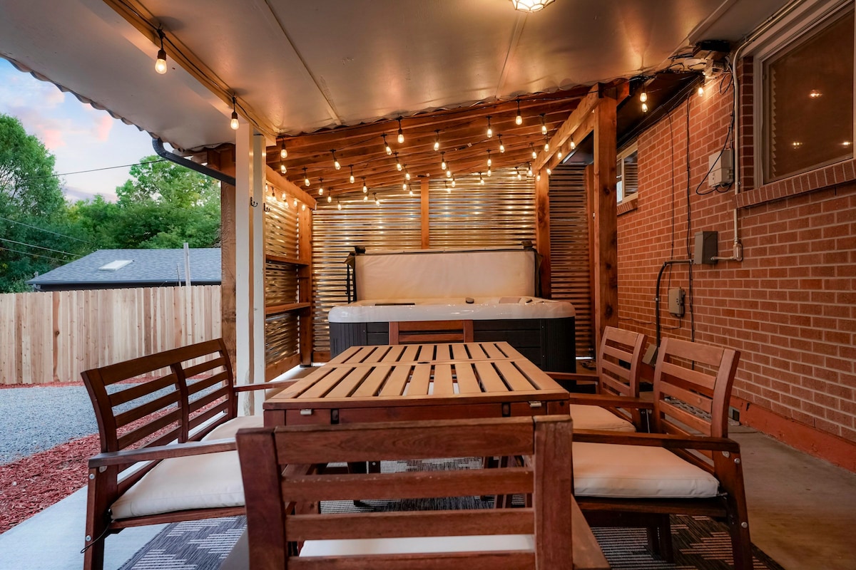 A covered patio area is shown, equipped with a wooden dining table and eight chairs, creating a welcoming space for gatherings. String lights hang above, providing soft illumination. A hot tub sits in the corner, inviting relaxation. The backdrop features a fenced backyard, enhancing privacy.