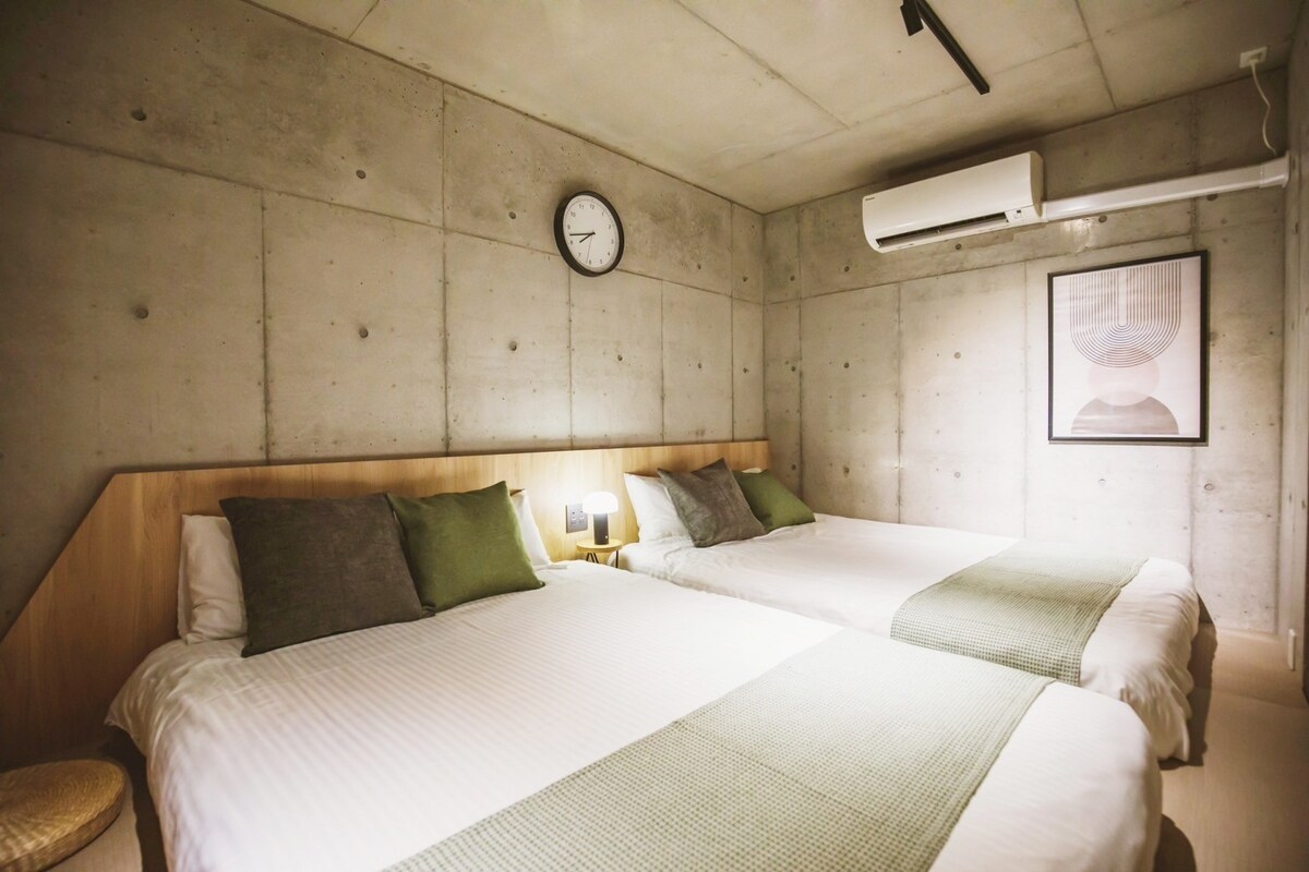 A minimalist bedroom features two spacious beds, each adorned with soft green accents. The concrete walls create a modern ambiance, complemented by a round wall clock and framed artwork. Natural light is filtered through a window, while an air conditioning unit is mounted above.