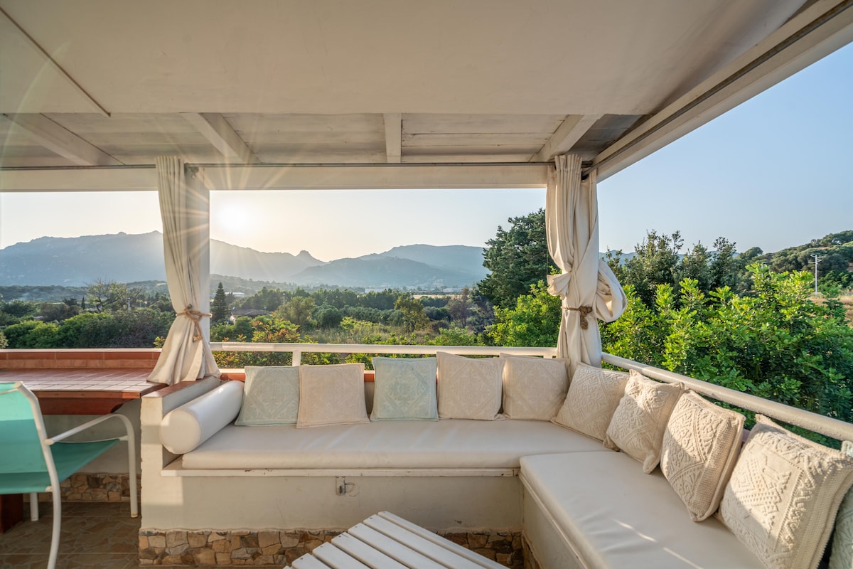 A spacious outdoor terrace features a built-in corner sofa with soft cushions, offering a panoramic view of surrounding hills and greenery. Natural light filters through the open space, creating a serene atmosphere ideal for relaxation or outdoor dining.