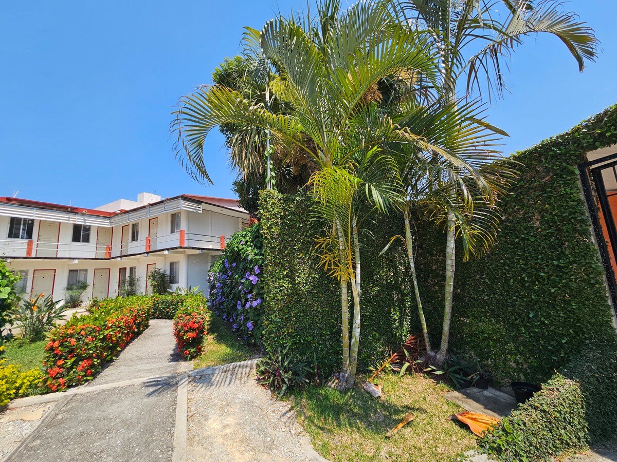 A well-maintained outdoor area is surrounded by lush greenery and colorful flower beds, providing a welcoming entrance. The pathway leads to a building with multiple entrances, which is situated against a clear blue sky.