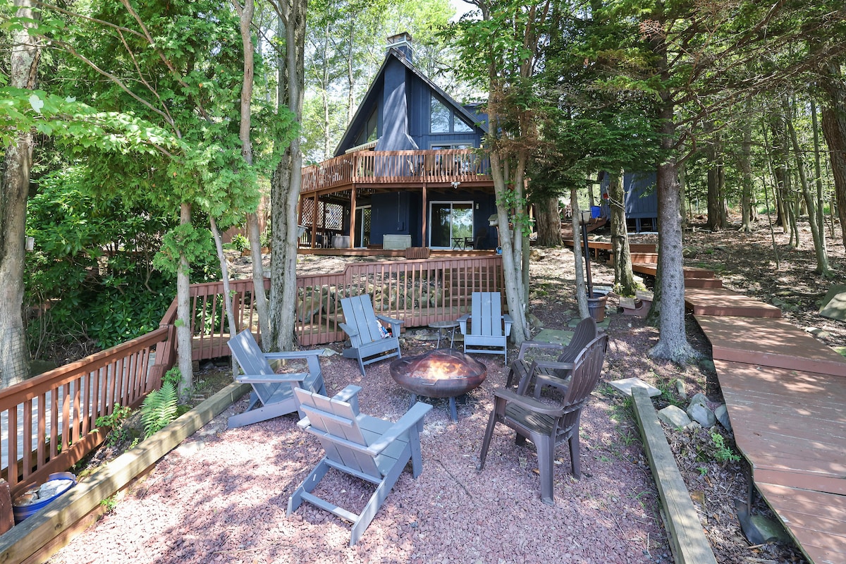 A cozy outdoor seating area is surrounded by trees. Five chairs, crafted from a combination of wood and plastic, are arranged around a fire pit. The cabin, featuring a modern A-frame design with large windows, stands in the background, offering a serene lakeside view.