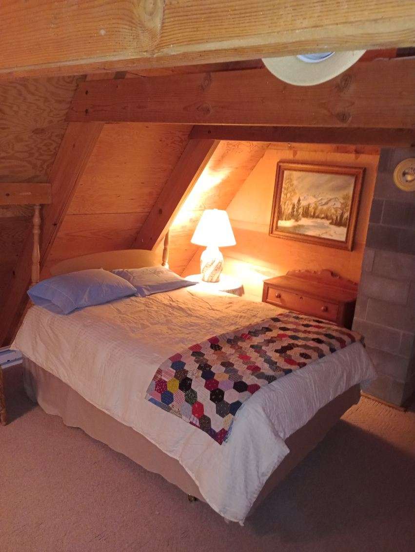 An attic-style bedroom features a cozy sleeping area with a spacious bed covered in a light quilt and an additional colorful throw. A bedside table holds a lamp, providing soft illumination. Wooden beams create a warm environment, complemented by a serene landscape painting on the wall.