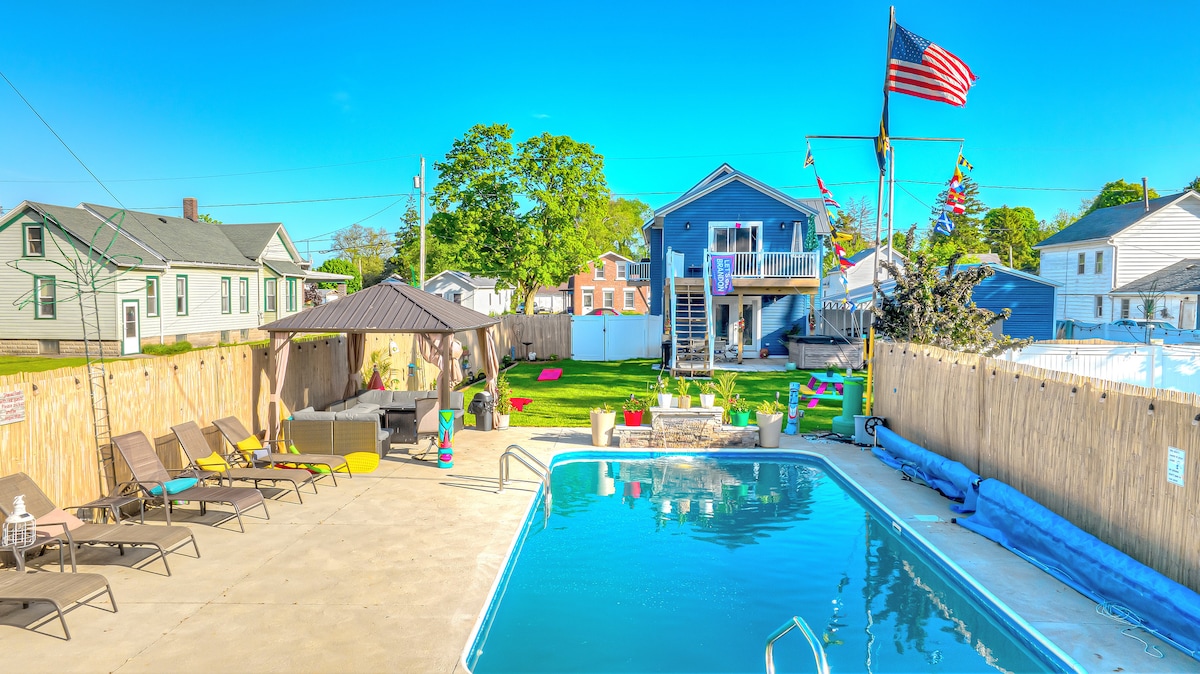 An inviting backyard features a heated, in-ground saltwater pool surrounded by a sun deck with lounge chairs. A gazebo provides shade, and a flag proudly flies above. The well-maintained yard extends with colorful seating areas, creating a perfect space for relaxation and enjoyment.