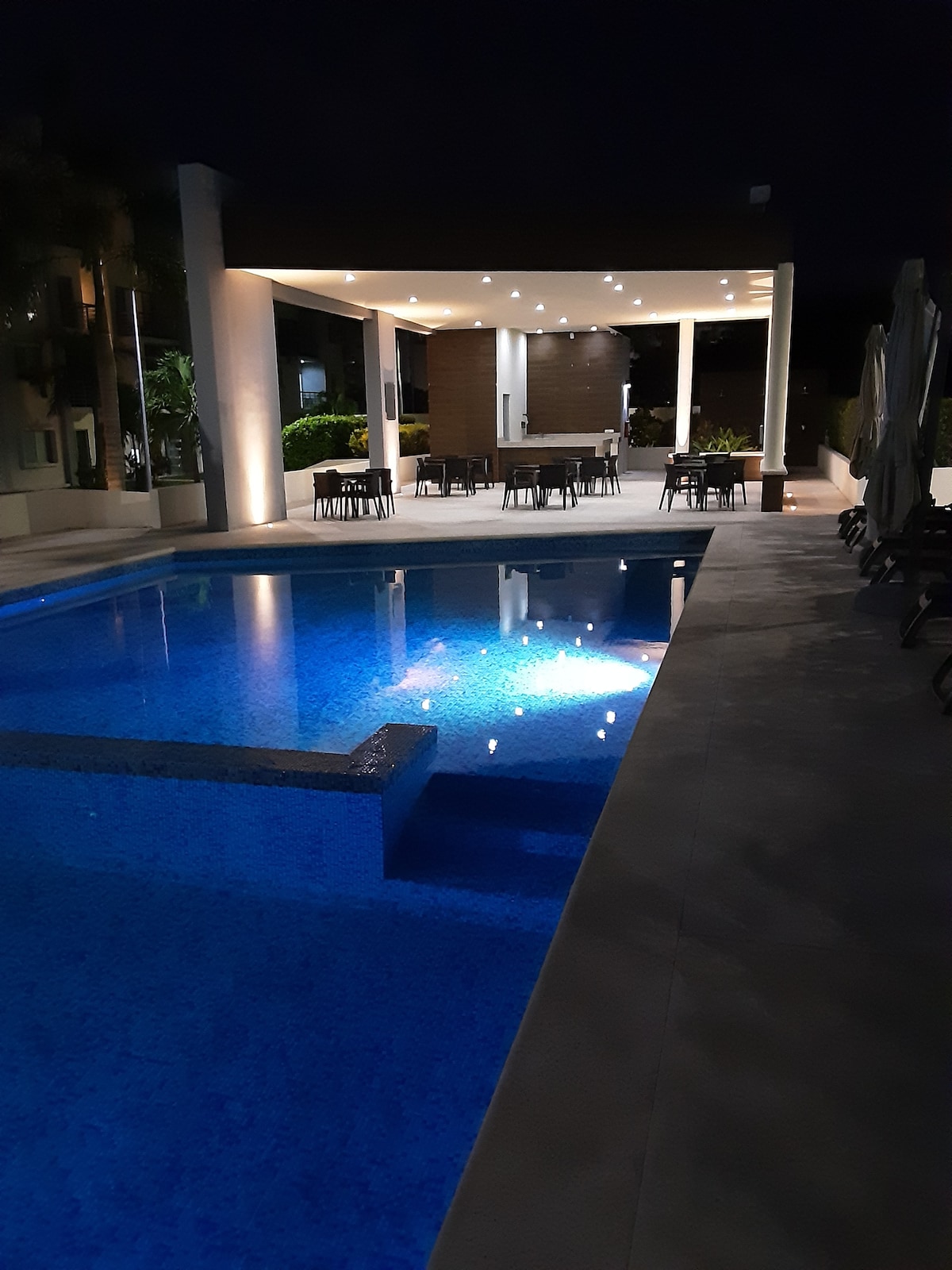 The serene pool area features clear blue water, which is softly illuminated by underwater lights. A spacious deck adjacent to the pool is furnished with tables and chairs, while a covered lounge area in the background offers additional seating and dining options.