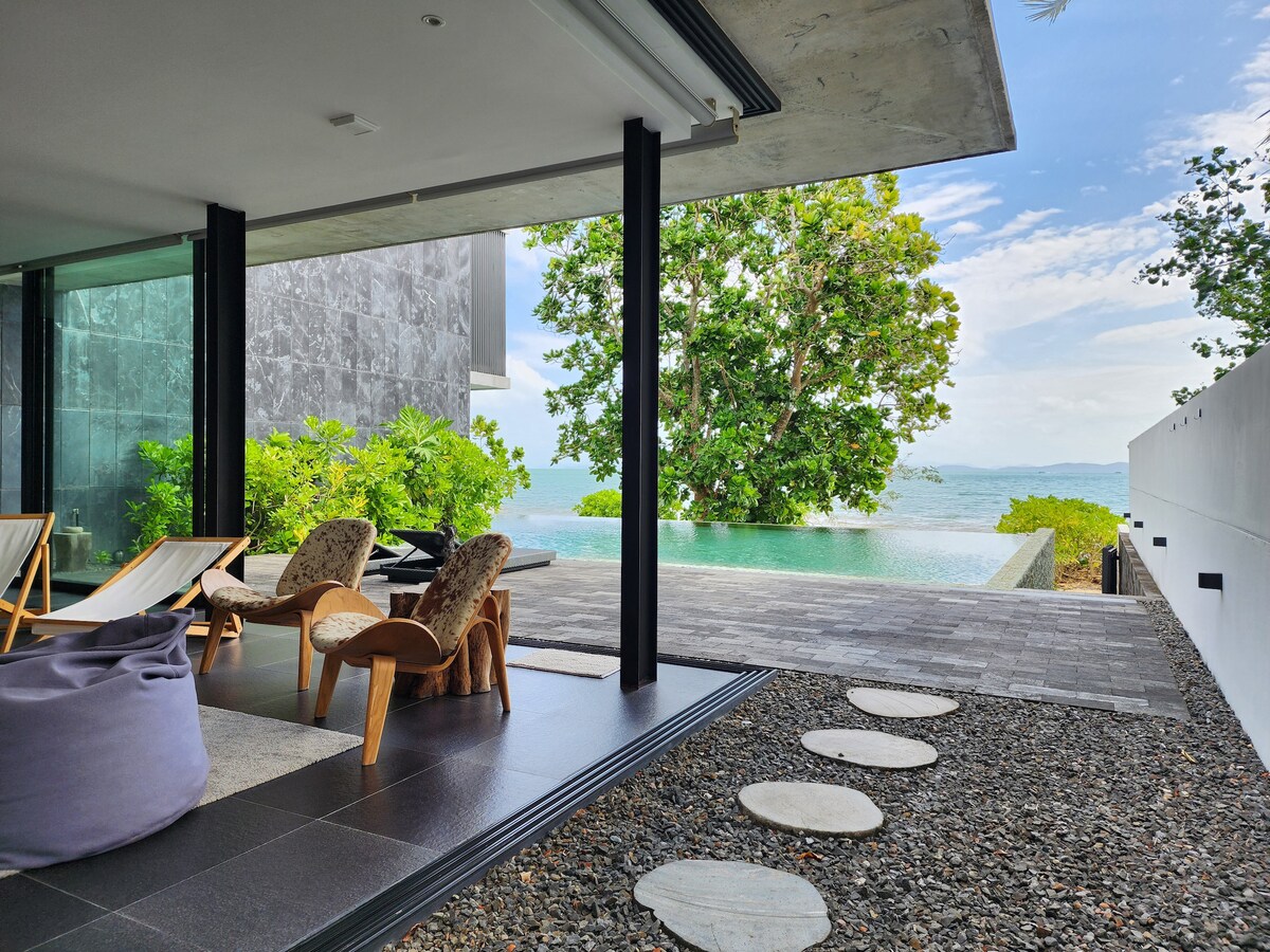 A spacious outdoor seating area is visible, featuring comfortable chairs and loungers placed on a stone and tile floor. A striking view of the ocean is framed by lush greenery, while an infinity pool extends toward the water's edge, creating a serene backdrop.